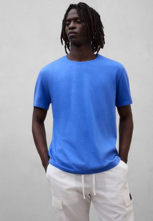 Mann mit dunkler Haut und Dreadlocks trägt ein blaues kurzärmeliges Hemd und weiße Cargohosen, steht mit den Händen in den Taschen vor einem einfarbigen Hintergrund.