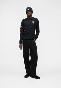 Μαύρο μπλουζάκι με μακριά μανίκια και πορτοκαλί σχέδια στα μανίκια, συνδυασμένο με μαύρο παντελόνι σε άνετη γραμμή και μαύρο σκουφί. Μαύρα παπούτσια ολοκληρώνουν την εμφάνιση.