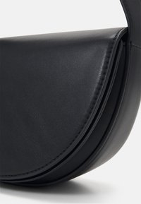 Gros plan sur un sac bandoulière en cuir noir avec rabat arrondi et détail de couture visible sur surface lisse.