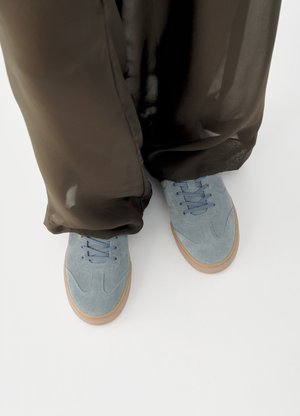 Svetlomodré semišové tenisky s gumovou podrážkou nosené s priesvitnými čiernymi širokými nohavicami na bielom pozadí.