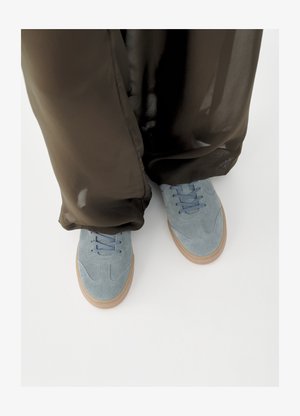 Baskets en daim bleu clair avec semelles en gomme portées avec un pantalon large transparent noir sur fond blanc.