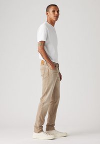 Uns jeans bege claro feitos de denim com um corte slim, combinados com uma t-shirt branca de algodão simples e ténis brancos, de pé contra um fundo neutro.