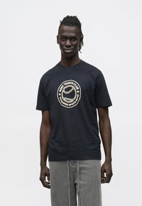 Tumesinine puuvillane T-särk ümara kaelusega, millel on rinnal keskel kreemikas logo. Lühikesed varrukad ja lõdvestunud istuvus.