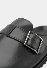 Μαύρο δερμάτινο σανδάλι τύπου slide με φαρδιά λουρά και ασημί ορθογώνια αγκράφα. Ομαλή υφή και διαμορφωμένο σχήμα, μινιμαλιστικός σχεδιασμός.