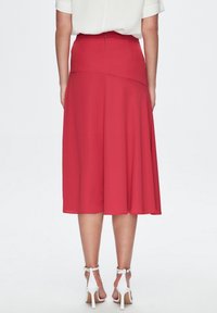 Falda roja de longitud a la rodilla con diseño acampanado, tela suave y cremallera oculta en la parte posterior. Llevada con sandalias de tacón alto blancas.