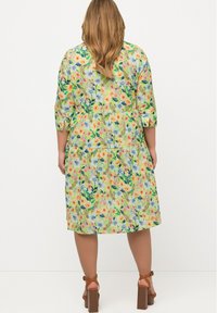 Vestido floral en verde claro con mangas de tres cuartos, una falda de volantes y un vibrante patrón de flores y hojas. Tacones gruesos marrones.