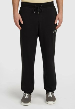 Mann mit schwarzen Jogginghosen mit Kordelzug in der Taille und grauen Turnschuhen steht vor einem schlichten Hintergrund.