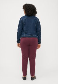 Ženska s kodrastimi lasmi, oblečena v denim jakno in burgundske hlače, stoji s hrbtom proti kameri na enobarvni beli podlagi.