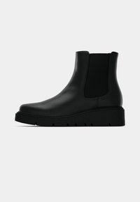 Μαύρο δερμάτινο μποτάκι μέχρι τον αστράγαλο με ελαστικές πλευρικές πάνελ, επίπεδη υφή σόλας και μινιμαλιστικό σχέδιο. Διαθέτει στρογγυλή μύτη και κομψή σιλουέτα.