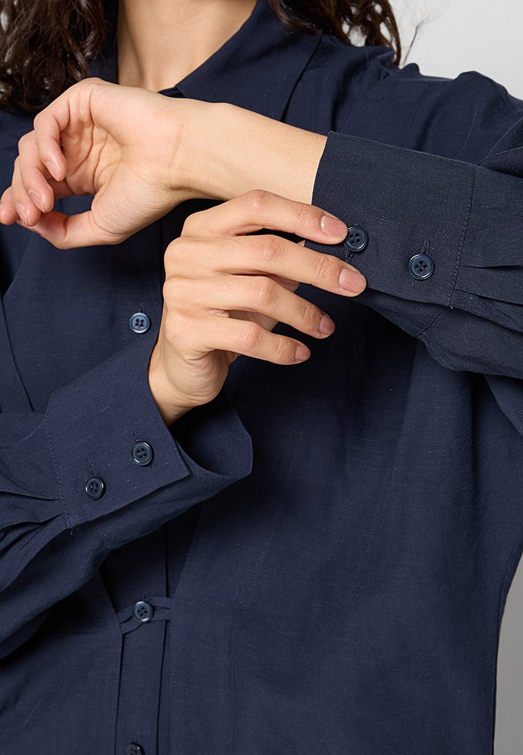 Camicia azzurra con bottoni, featuring un colletto classico, maniche a pieghe e due bottoni scuri sui polsini. Tessuto liscio e strutturato.