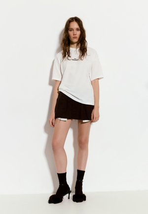 Weißes, oversize T-Shirt mit schwarzem Text, kombiniert mit einem braunen, gefalteten Minirock. Schwarze Mules mit Absätzen und Furr-Akzenten sowie knielangen Socken.