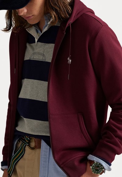 Sweat à zip bordeaux avec une poche avant, porté par-dessus un polo rayé gris et marine. Accentué par un petit logo et associé à un pantalon beige.