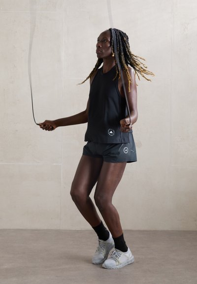 Black sleeveless top and black shorts, with a logo detail. Athlete skipping rope, wearing gray sneakers and black socks against a neutral backdrop.