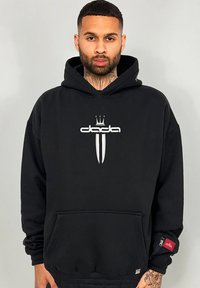 Schwarzer Hoodie aus Baumwollmischung, mit einem weißen Logo mit einer Krone und zwei vertikalen Linien. Enthält eine Fronttasche und einen Reißverschluss an den Bündchen.