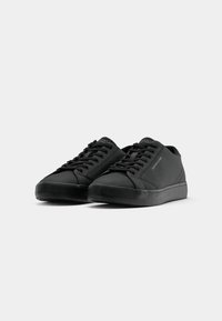 Paire de baskets basses noires avec lacets, avec un marquage subtil sur le côté et la languette, sur un fond clair uni.