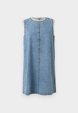 Femme portant une robe en jean bleu clair sans manches avec des bords festonnés, portant un sac à épaule crème et des sandales noires, debout.