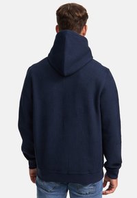 Marineblå hettegenser med teksturert stoff og ribbestrikket hette. Har et mønster av vertikale striper. Lange ermer og en tettsittende midje.