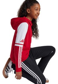 Rode hoodie met witte mouwen en drie witte strepen op de armen, gecombineerd met zwarte broek met witte strepen. Witte sneakers met bruine zolen.