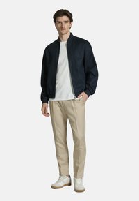 Man wearing navy bomber jacket, white t-shirt, beige pants, and white sneakers, standing with one hand in pocket, neutral background.