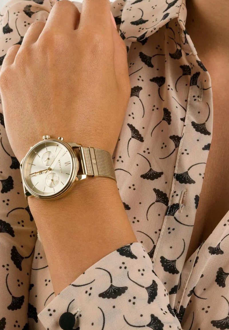 Montre dorée avec un bracelet en maille, cadran rond présentant des détails argentés et blancs, positionnée sur une main portant une blouse à motifs avec des accents noirs.