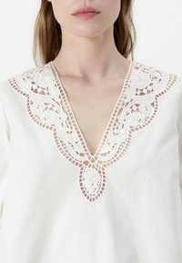 Blouse blanche ornée de détails en dentelle délicate le long d'un décolleté en V profond, avec des motifs floraux et des accents découpés au niveau du col.