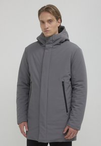 Cappotto imbottito grigio con colletto alto e cappuccio. Presenta una tasca laterale con zip e una silhouette slanciata. Texture liscia.