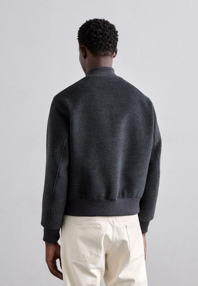 Chaqueta bomber gris con un acabado texturizado, cuello y puños acanalados, combinada con pantalones color crema, vista desde la espalda.