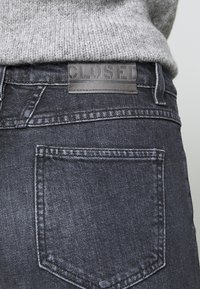 Pull gris en maille rentré dans un jean en denim foncé montrant un patch en cuir avec le mot "CLOSED" au-dessus de la poche arrière.