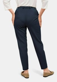 Marineblauwe op maat gemaakte broeken met een hoge taille, plooien aan de voorkant en taps toelopende pijpen, gecombineerd met tan instappers. De stof ziet er glad uit.