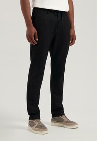 Schwarze Jogginghosen aus weichem Material mit einem Kordelzugbund, seitlichen Taschen und einer taillierten Passform, kombiniert mit grauen Turnschuhen.