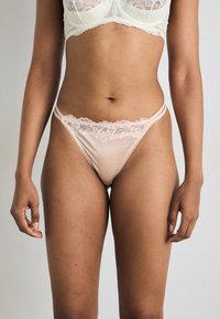 Lichtroze satijnen string met delicate kantafwerking, dunne bandjes en fijne stiksels. Gladde textuur en een ontwerp met minimale bedekking.