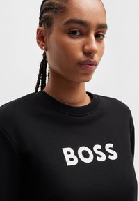 Black sweatshirt with a crew neckline and textured fabric. Features bold white "BOSS" text on the front. Close-up of a person wearing it.