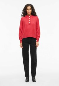 Punane soonikkoes kampsun kõrge kraega ja kolme ees oleva nupuga, mida kannavad mustad laia säärega püksid ja mustad kontsakingad.