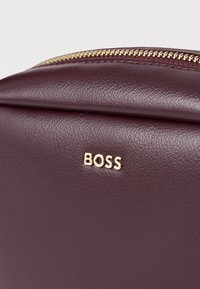 Μπορντό δερμάτινη τσάντα με υφή, με χρυσό λογότυπο "BOSS" και χρυσό φερμουάρ στην κορυφή.