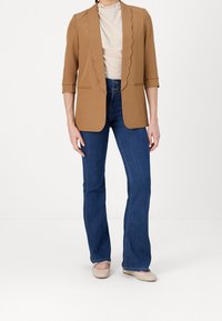 Personne portant un blazer marron à bord festonné, un haut beige, un jean bleu taille haute et des chaussures plates beiges, debout devant un fond blanc.
