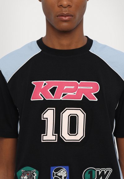 Camiseta negra con el logo "KPR" en rosa, el número "10" en rosa pálido y acentos azules en los hombros. Presenta parches decorativos en la parte inferior.
