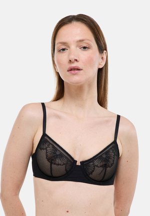 Femme aux cheveux bruns lisses porte un soutien-gorge en dentelle noire transparente avec des motifs floraux, posant devant un fond clair uni.