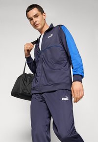Moški v temno modrem Puma trenirka drži črn športni nahrbtnik na rami pred svetlo ozadjem.
