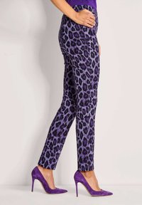 Vrouw in paarse broek met luipaardprint en bijpassende paarse hoge hakken, staand in zijaanzicht met hand op de heup.