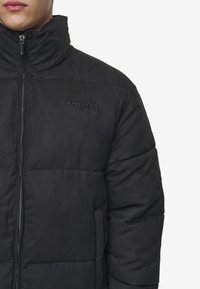 Svart puffjacka med hög krage, dragkedja och präglad logotyp. Har sydda paneler och en slät yta.