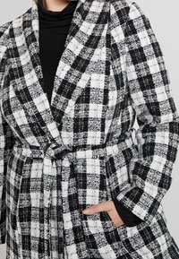 Blazer a cuadros en blanco y negro hecho de tejido texturizado, con cintura ceñida por un cinturón, cuello con muescas y bolsillos laterales.