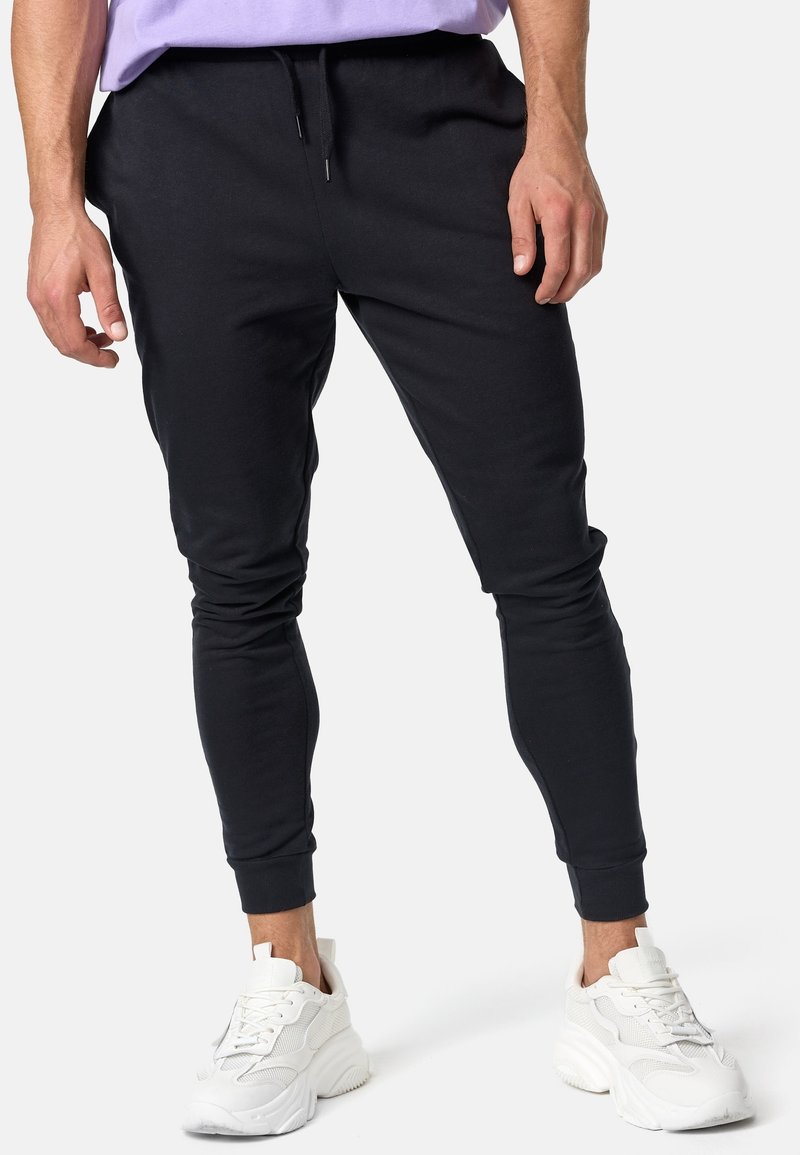 Hombre vistiendo pantalones jogger ajustados negros y zapatillas deportivas gruesas blancas, de pie contra un fondo claro y liso.