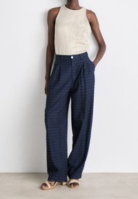 Beige mouwloos topje gecombineerd met marineblauwe, wijde broek met subtiele strepen en voorplooien. Model staat op een neutrale ondergrond.