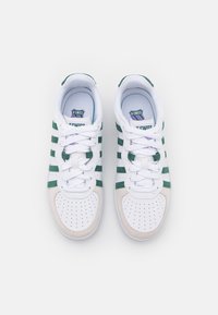 Bílé tenisky se zelenými semišovými pruhy, texturovaným bílým síťovaným jazykem, kulatou špičkou a perforováním. Fialové logo na jazyku.