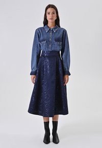Denim shirt met twee voorzakken, gecombineerd met een marineblauwe midi-jurk met bloemenpatroon. Model draagt zwarte hoge laarzen.