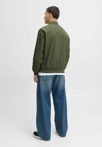 Veste bomber vert olive avec un col et des poignets côtelés, associée à un jean en denim bleu à jambes larges et des chaussures noires. Texture lisse, design minimaliste.