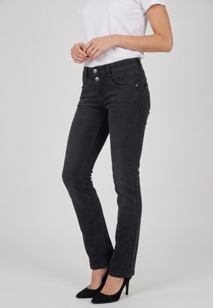 Donna con jeans aderenti neri, maglietta bianca a maniche corte e scarpe nere con tacchi alti, in piedi davanti a uno sfondo uniforme.