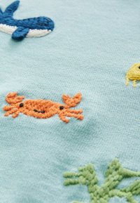 Svetlo modra tkanina vsebuje barvite vezenine morskega življenja: modra kita, oranžnega raka, rumeno morsko zvezdo in zelene detajle morske trave.