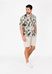 Camisa de manga corta beige con cuello y patrón de hojas verdes, combinada con pantalones cortos beige claros y zapatillas blancas. Tela suave y corte casual.