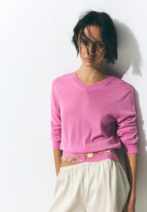 Femme aux cheveux courts et foncés porte un pull rose et un pantalon crème, accessoirisée d'une ceinture circulaire dorée, debout devant un mur blanc.